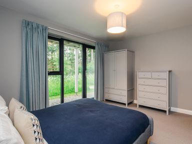 A bedroom with a bed with blue bedding a wardrobe and chest of drawers next to large glass doors with curtains at Wildcat Lodge in Feshiebridge near Aviemore
