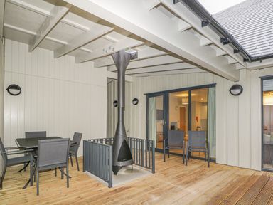 An outdoor covered patio with a metal fireplace surrounded by a railing a table with four chairs two armchairs and glass doors leading inside at Golden Eagle Lodge in Aviemore