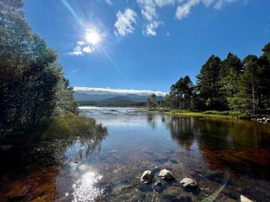 A river with trees on both sides and mountains in the background under a sunny sky at Golden Eagle Lodge in Aviemore