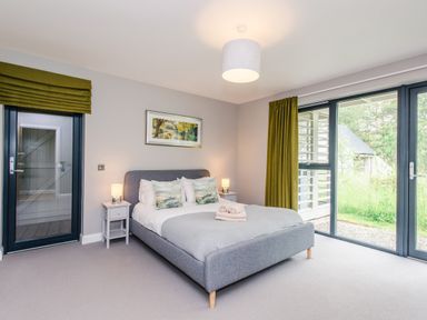 A bedroom with a grey bed a pair of rolled towels bedside tables with lamps and large glass doors with green curtains at Golden Eagle Lodge in Aviemore