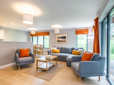 A living room with grey sofas and armchairs with orange cushions and curtains a wooden coffee table and dining table with chairs at Red Deer Lodge in Feshiebridge near Aviemore
