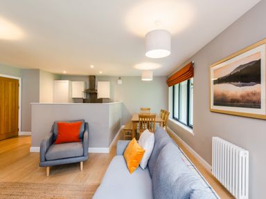 A living room with a gray sofa and armchair with orange pillows and a dining table near kitchen at Red Deer Lodge in Feshiebridge near Aviemore