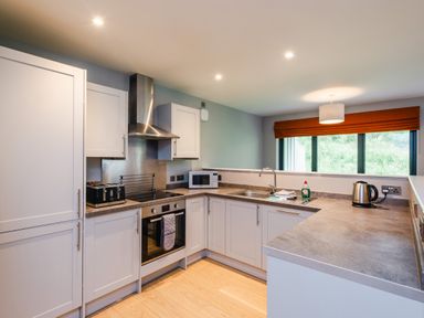 A kitchen with white cabinets a stove microwave sink kettle toaster and a window with an orange blind at Red Deer Lodge in Feshiebridge near Aviemore