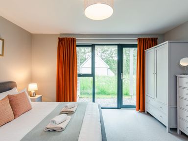 A bedroom with a bed, bedside table and lamp, wardrobe, chest of drawers, and glass doors with orange curtains at Red Deer Lodge in Feshiebridge near Aviemore