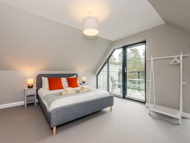A bedroom with a gray upholstered bed with orange pillows bedside tables with lamps a balcony door and a white clothes rack at Red Deer Lodge in Feshiebridge near Aviemore