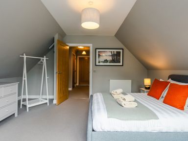 A bedroom with a bed with orange pillows and folded towels a white dresser and a white clothes rack with an open wooden door and a framed picture on the wall at Red Deer Lodge in Feshiebridge near Aviemore