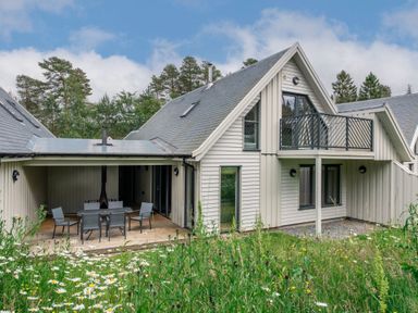 An exterior view of a two story house with a covered patio and balcony surrounded by grass and trees at Red Deer Lodge in Feshiebridge near Aviemore
