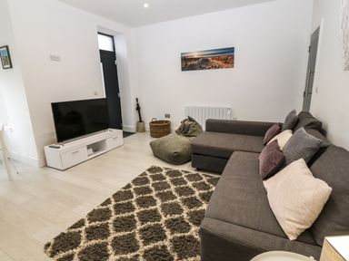 A living room with a gray sectional sofa, a patterned rug, a bean bag, a white TV stand with a television, and wall art at Arlanfor in Rhosneigr
