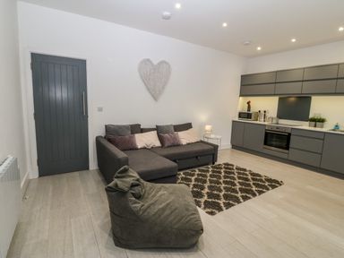 A living room with a gray sofa, a bean bag chair, a patterned rug, and a gray kitchen area at Arlanfor in Rhosneigr