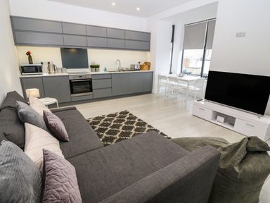 A living room with a grey sofa and cushions rug and kitchen with grey cabinets and dining table in the corner at Arlanfor in Rhosneigr