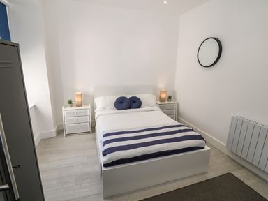 A bedroom with a white bed with blue pillows and a blue striped blanket two white nightstands with lamps and small plants a black round mirror on the wall at Arlanfor in Rhosneigr