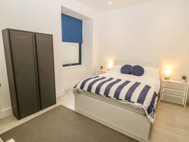 A bedroom with a white bed striped blanket blue cushions two bedside tables with lamps and plants a grey wardrobe and a blue window blind at Arlanfor in Rhosneigr