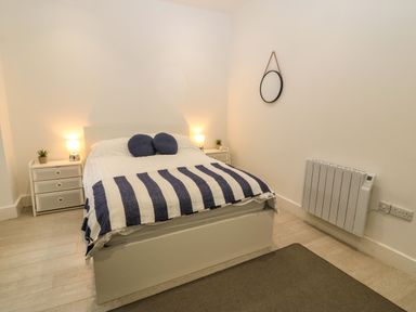 A bedroom with a bed with striped blanket and two pillows two bedside tables with lamps and plants a round mirror on the wall and a heater on the floor at Arlanfor in Rhosneigr