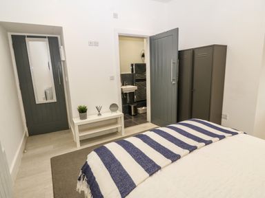 A bedroom with a bed covered in a striped blanket two gray doors a white bench with plant and reed diffuser and view into a bathroom at Arlanfor in Rhosneigr