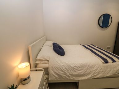 A bedroom with a white bed and bedding a round blue pillow a striped blanket a white nightstand with a lamp and a small plant and a round mirror on the wall at Arlanfor in Rhosneigr