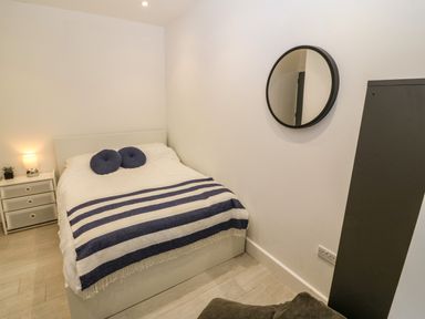 A bedroom with a bed with white bedding blue cushions and a striped blanket a bedside table with a lamp and plant a round mirror and a tall dark cabinet at Arlanfor in Rhosneigr