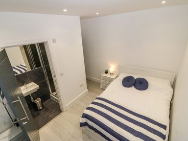 A bedroom with a double bed with blue round cushions and a striped blanket next to a small white bedside table with a lamp and a plant and an open door leading to a bathroom at Arlanfor in Rhosneigr