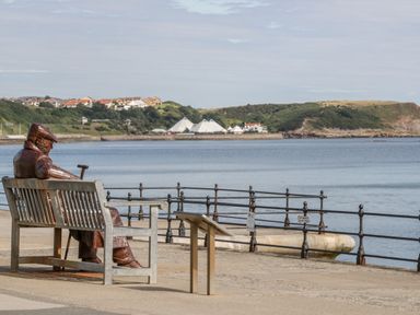 A statue of a man sitting on a bench looking out over water with houses and hills in the background at Sanderling in Scalby