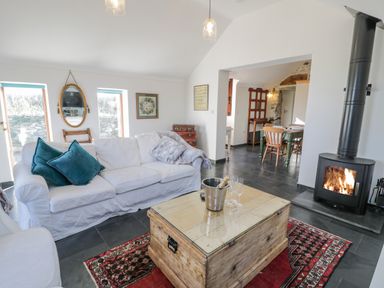 A living room with a white sofa and cushions a wooden chest used as a coffee table with a champagne bucket and glasses a fireplace and a dining area visible through an archway at Cwch Gwenyn in Llanfachraeth