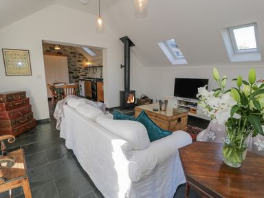 A living room with a white sofa and teal cushions a wood-burning stove a wooden coffee table and television next to a kitchen with wooden cabinets and dining table at Cwch Gwenyn in Llanfachraeth