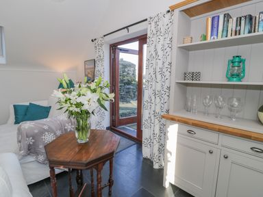 A sitting area with a wooden table holding a vase of white flowers next to a white sofa with cushions and a blanket near a glass door and a white cabinet with books and glassware at Cwch Gwenyn in Llanfachraeth