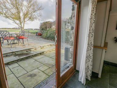 A wooden door opening to a stone patio with metal chairs and table with red cushions at Cwch Gwenyn in Llanfachraeth