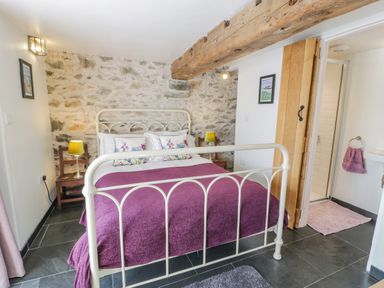 A bedroom with a white metal bed frame, purple bedding, wooden chairs with lamps, stone wall, wooden beam on ceiling, and a door leading to a bathroom at Cwch Gwenyn in Llanfachraeth