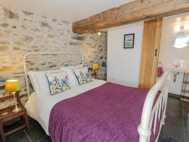 A bedroom with a white metal bed frame purple blanket floral pillows exposed stone wall wooden beam bedside tables with lamps and a small sink area at Cwch Gwenyn in Llanfachraeth