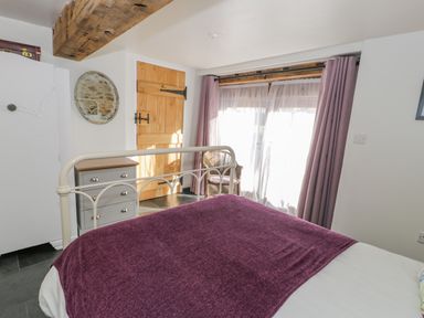 A bedroom with a white metal bed frame purple blanket a wooden door a wicker chair and gray chest of drawers at Cwch Gwenyn in Llanfachraeth