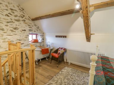 A bedroom with a bathtub beside a stone wall and wooden staircase at Cwch Gwenyn in Llanfachraeth
