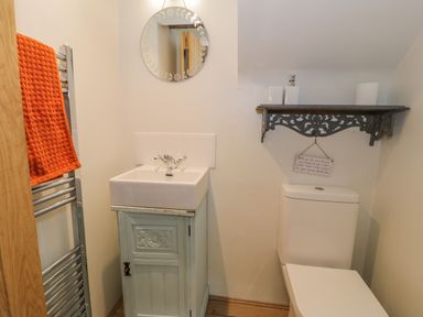 A small bathroom with a sink on a cabinet a round mirror a toilet a shelf with accessories and an orange towel on a heated rack at Cwch Gwenyn in Llanfachraeth