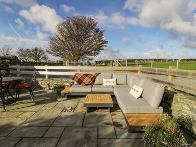 An outdoor seating area with a sectional sofa and table on stone paving surrounded by wooden fencing and stone walls with fields and trees in the background at Cwch Gwenyn in Llanfachraeth