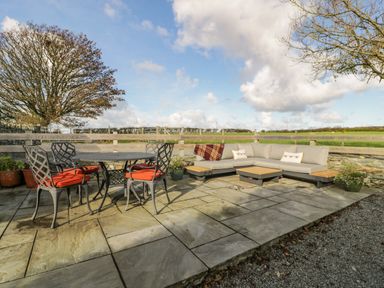 An outdoor patio with a round metal table and four chairs with red cushions and a corner sofa with beige cushions near a wooden fence in a rural setting at Cwch Gwenyn in Llanfachraeth