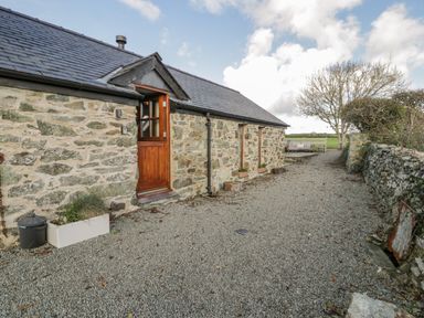 A stone cottage with a wooden door and two windows next to a gravel path with a seating area in a rural setting at Cwch Gwenyn in Llanfachraeth