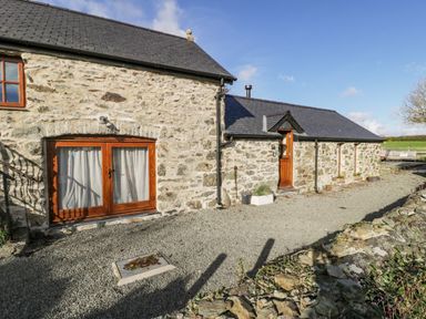 A stone cottage with wooden doors and windows on a gravel driveway at Cwch Gwenyn in Llanfachraeth