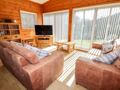 A living room with two brown sofas a wooden coffee table a TV on a wooden stand and large windows with vertical blinds at Watersview Lodge Rudyard near Leek