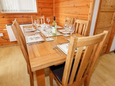 A wooden dining table with six chairs set with placemats glasses cutlery and a bottle in a room with wooden walls at Watersview Lodge in Rudyard near Leek
