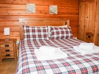 A bedroom with a wooden bed with plaid bedding two folded towels on the bed wooden nightstand with a lamp wooden wardrobe and two heart-shaped wall decorations at Watersview Lodge Rudyard near Leek