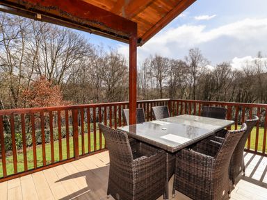An outdoor balcony with a glass table and six wicker chairs surrounded by wooden railing at Watersview Lodge in Rudyard near Leek