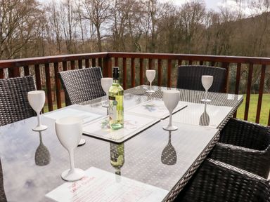 An outdoor patio with a glass table set with six white wine glasses and a bottle of white wine surrounded by wicker chairs at Watersview Lodge Rudyard near Leek