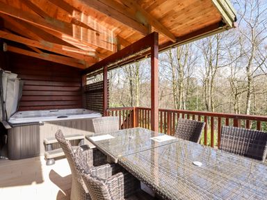 An outdoor covered wooden deck with a hot tub and a glass top table surrounded by wicker chairs at Watersview Lodge in Rudyard near Leek