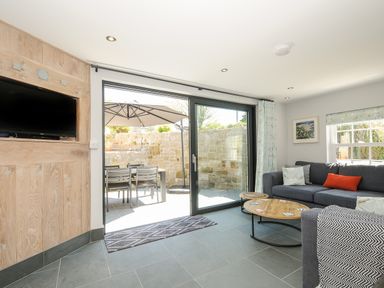 A living room with a gray sofa a coffee table a wall-mounted television and sliding glass doors leading to an outdoor dining area at Stargazy Cottage in Praa Sands