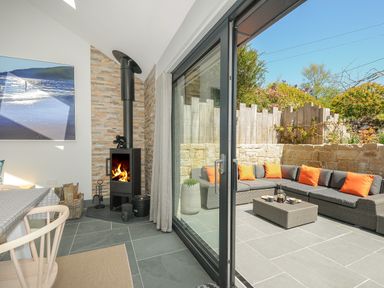 A living room with a wood burning stove next to sliding glass doors opening to a patio with sectional sofa with orange cushions at Stargazy Cottage in Praa Sands