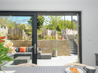 A patio with outdoor seating and orange cushions viewed through sliding glass doors with a table set with a croissant and cup in the foreground at Stargazy Cottage in Praa Sands