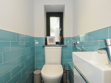 A small bathroom with a white toilet blue tiled walls a window above the toilet and a white sink with silver faucet at Stargazy Cottage in Praa Sands