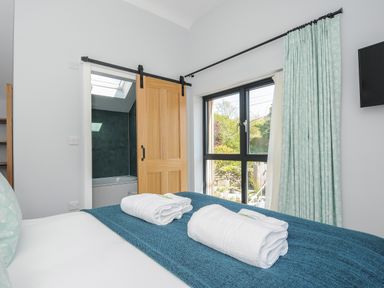 A bedroom with a bed with folded towels a wooden sliding door leading to a bathroom and a window with patterned curtains at Stargazy Cottage in Praa Sands