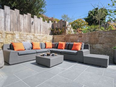 A gray outdoor sectional sofa with orange pillows on a tiled patio with stone and wood fencing at Stargazy Cottage in Praa Sands