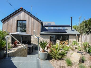 A garden with plants and rocks leading to a house with wooden and stone exterior and solar panels on the roof at Stargazy Cottage in Praa Sands