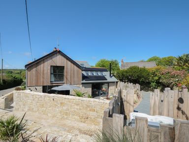 A wooden and stone house with solar panels a stone wall and a fenced patio with a hot tub at Stargazy Cottage in Praa Sands