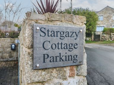 A stone wall with a wooden sign reading Stargazy Cottage Parking near a paved road and stone buildings at Praa Sands